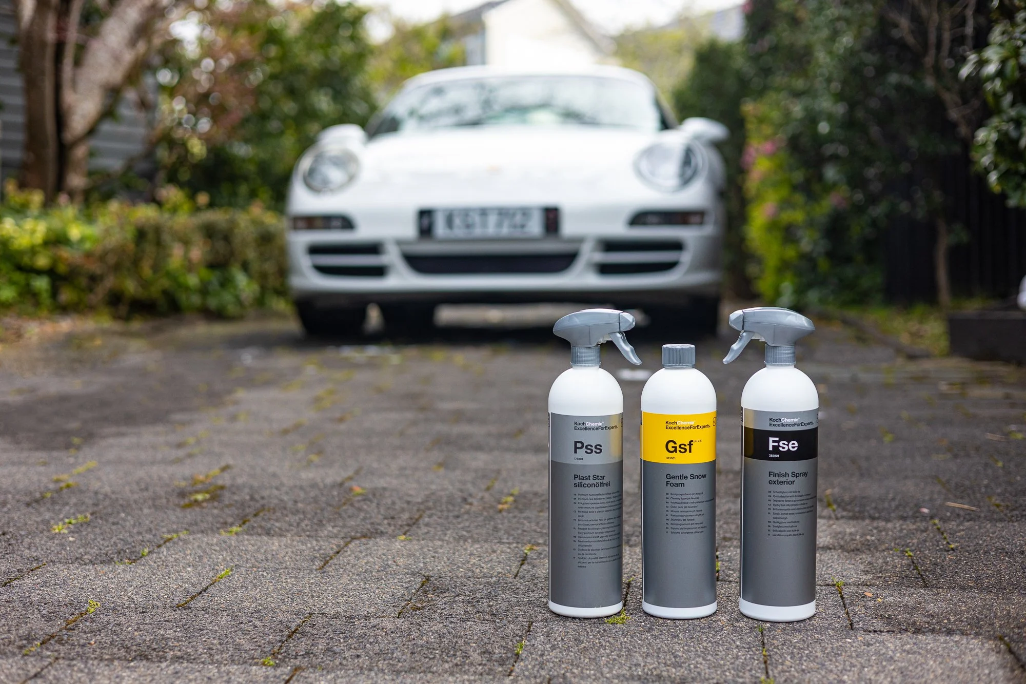 Three bottles of car detailing spray on a driveway with a white car in the background.