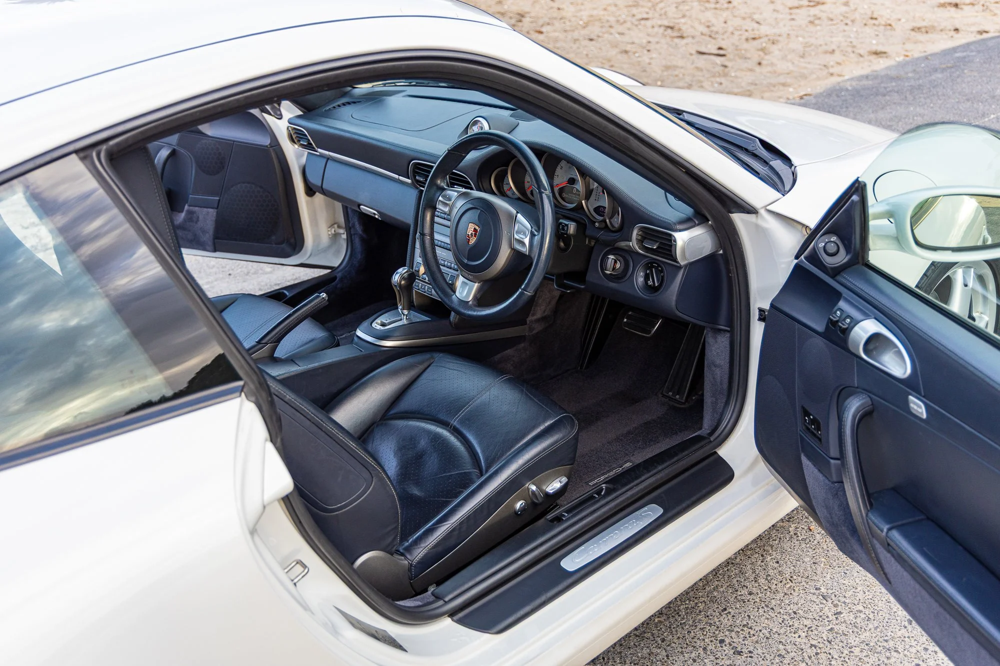 Interior view of a white Porsche sports car showing the steering wheel, dashboard, leather seats, and open driver-side door.