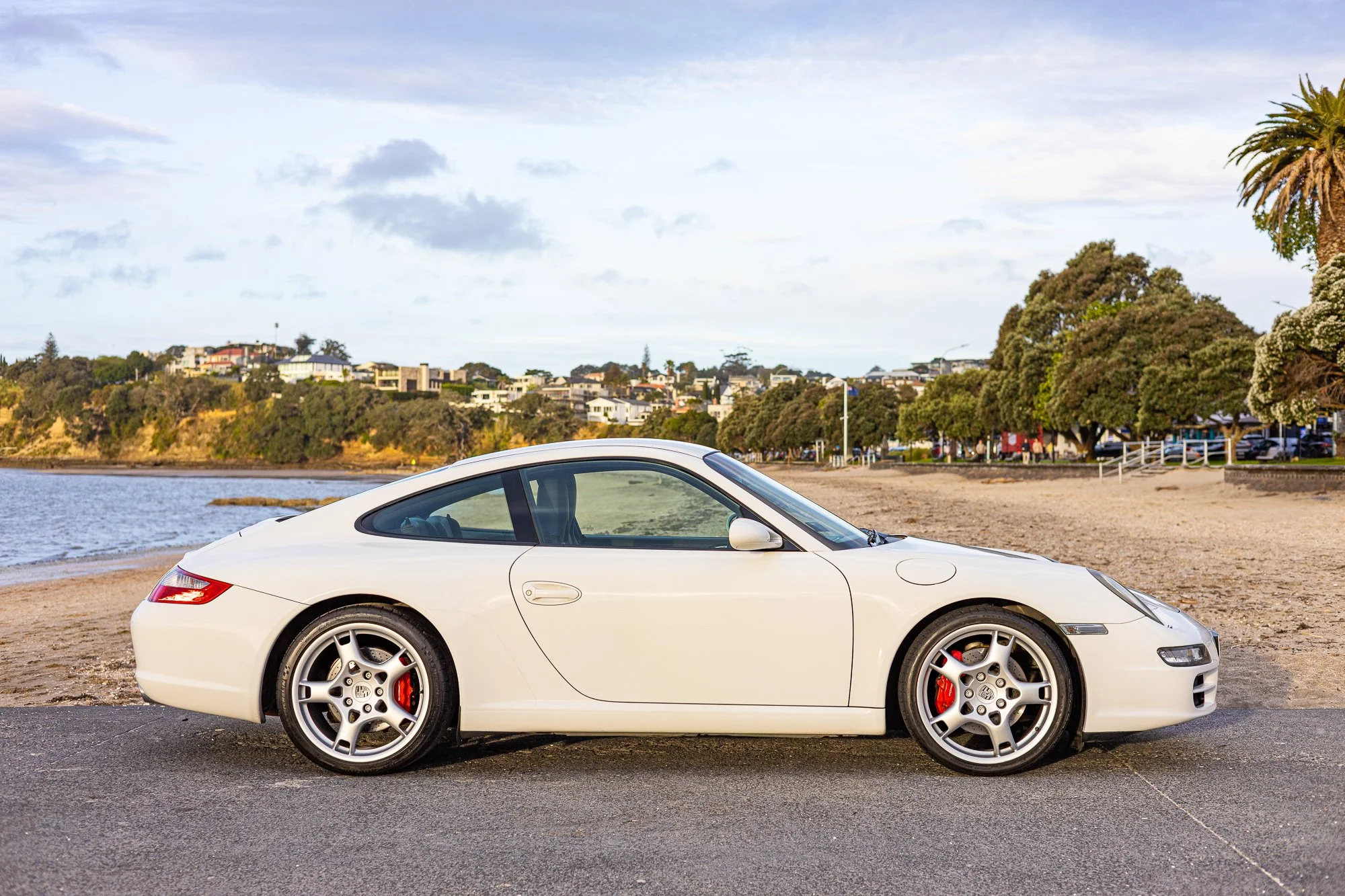 White Porsche sports car parked on a street near a beach with trees and buildings in the background