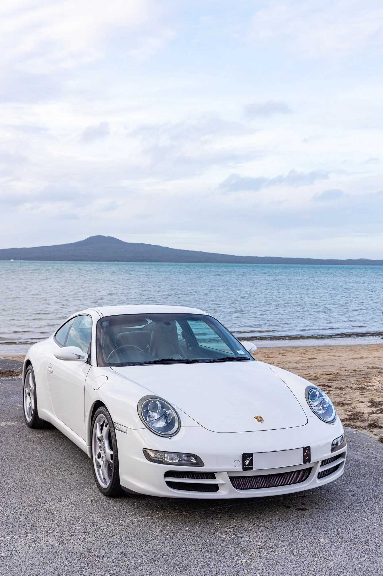 White Porsche sports car parked on a coastal road near the beach with ocean and distant hills in the background.