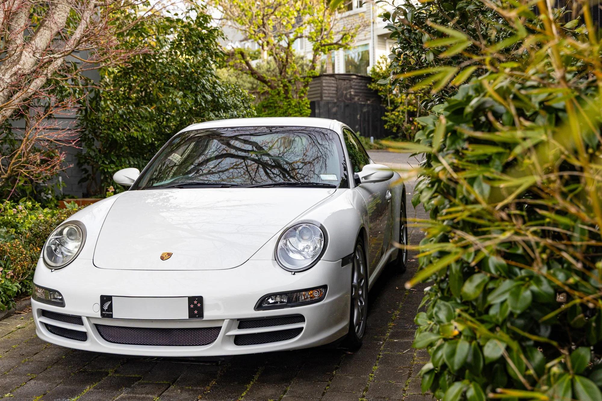 White Porsche sports car parked on a brick driveway, partially obscured by green bushes and trees in a residential area.