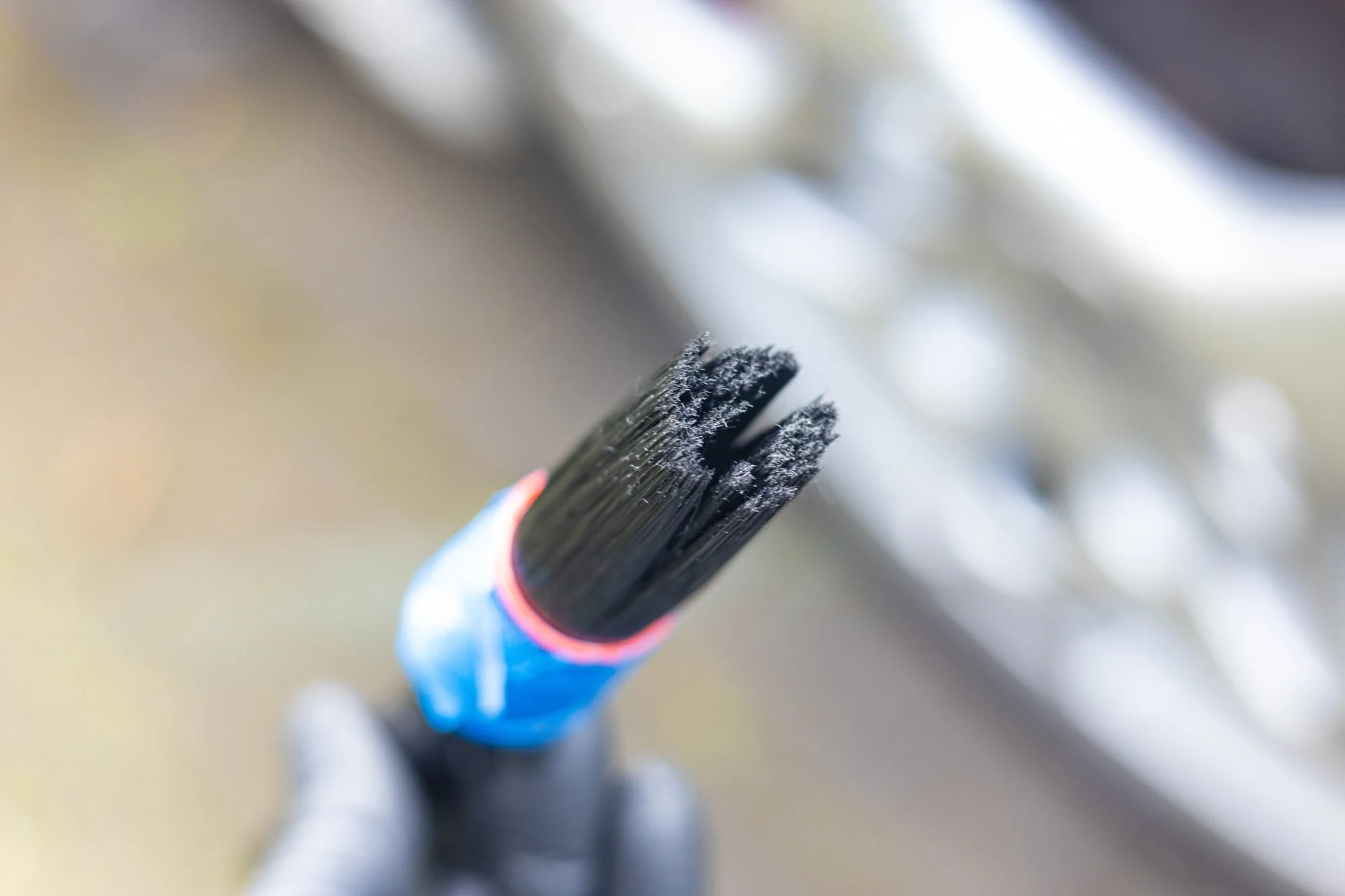 Close-up of a hand wearing a glove holding a paintbrush with black bristles covered in frost, outdoors with blurred background.