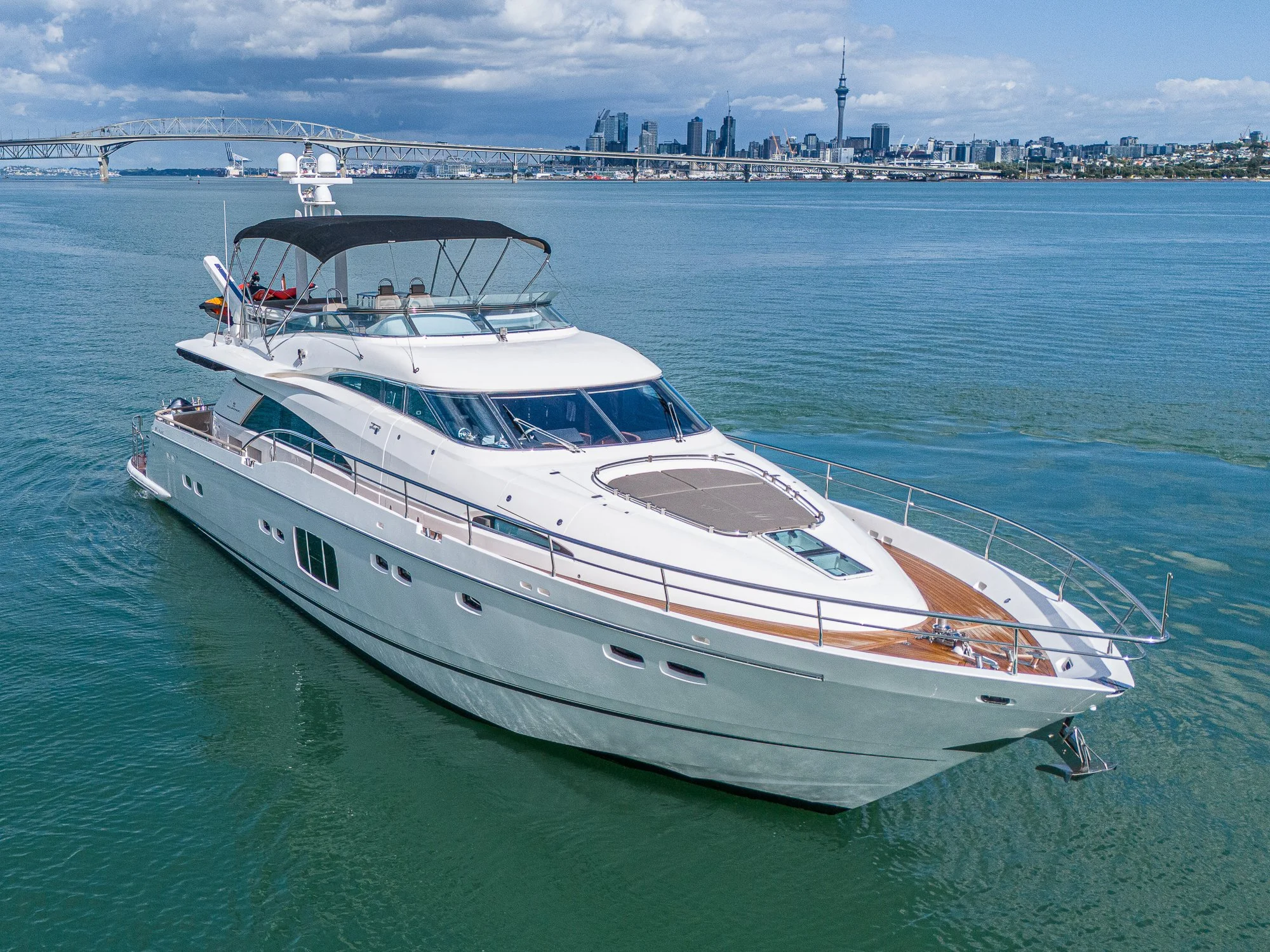 A large white yacht with wooden decking on the bow, sailing on calm water with a city skyline and bridge in the background.