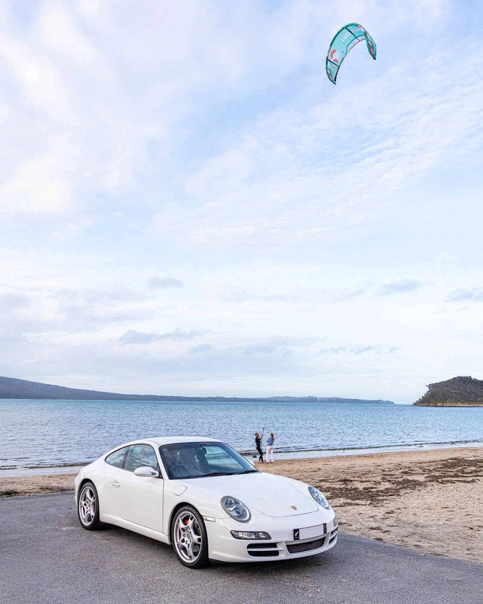 A white Porsche sports car parked near a beach with the ocean in the background. Two people are flying a kite on the beach.