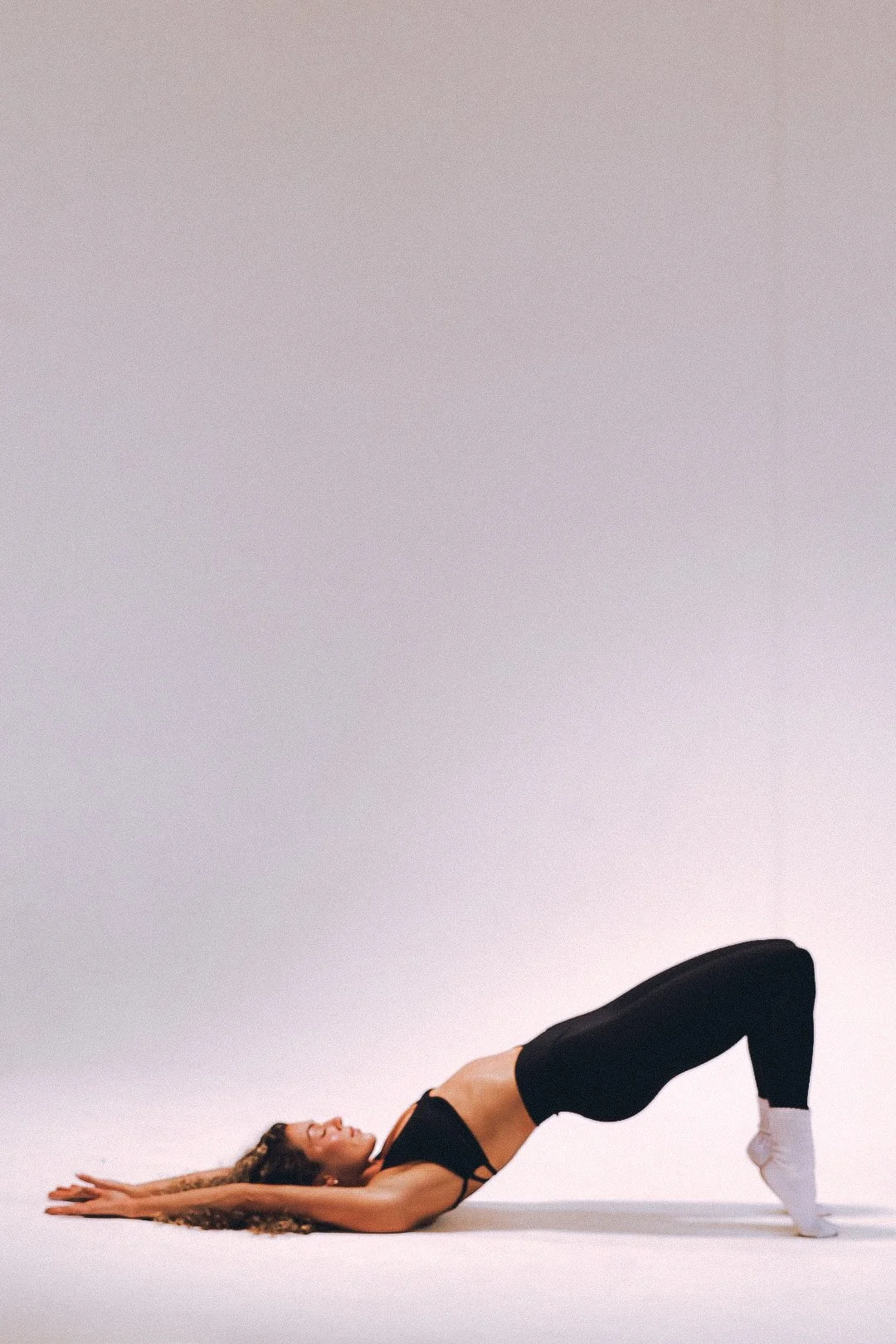 A woman lying on her back doing a leg lift exercise on a light-colored floor, wearing black workout clothes and white socks, against a plain light background.