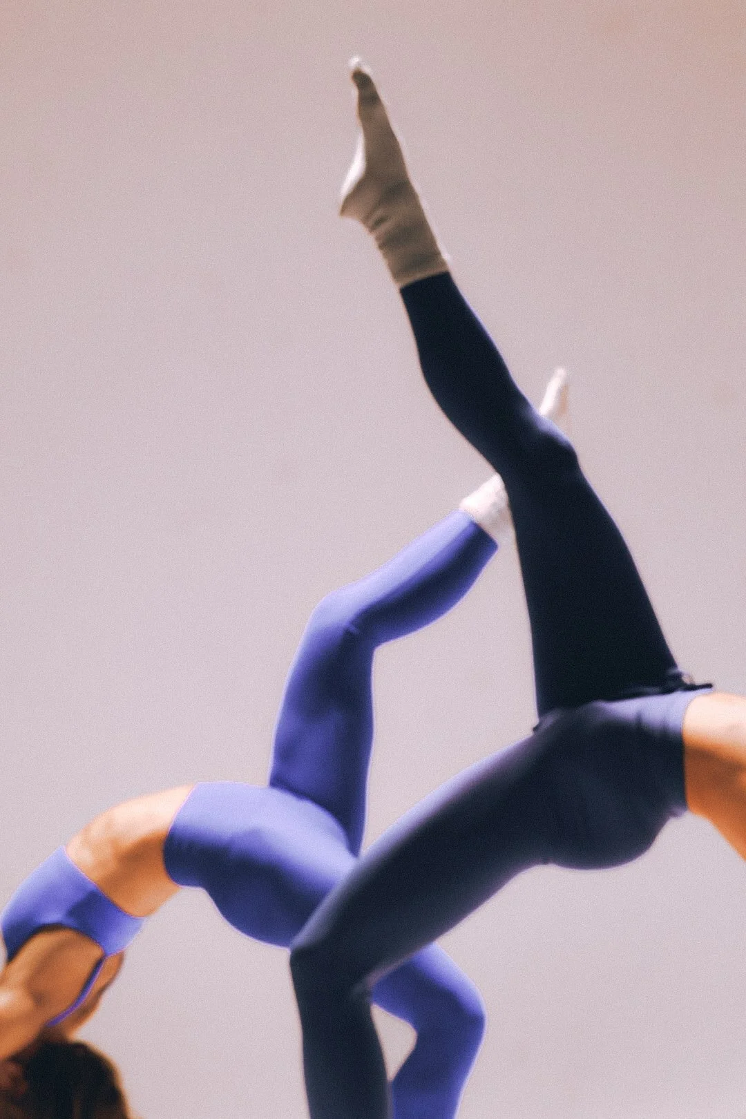Fitness studio, two people in a split pose with one leg extended upward and the other downward, wearing blue leggings and socks, against a plain background.
