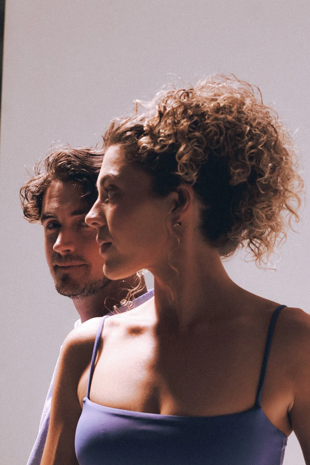 A man and a woman standing close together, with the woman in front and the man slightly behind her, both facing to the left against a plain, light-colored background.