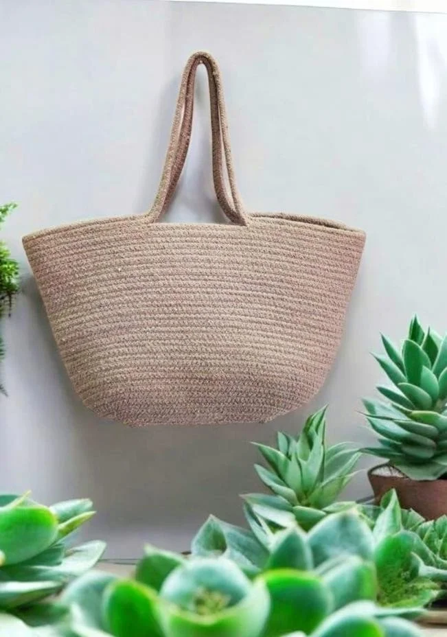 Beige woven tote bag hanging on a wall surrounded by green succulents.