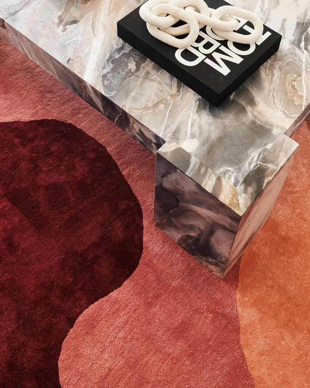 Close-up of a marble table with a decorative white chain and a black book with white text, next to a red area rug with darker red circle design.