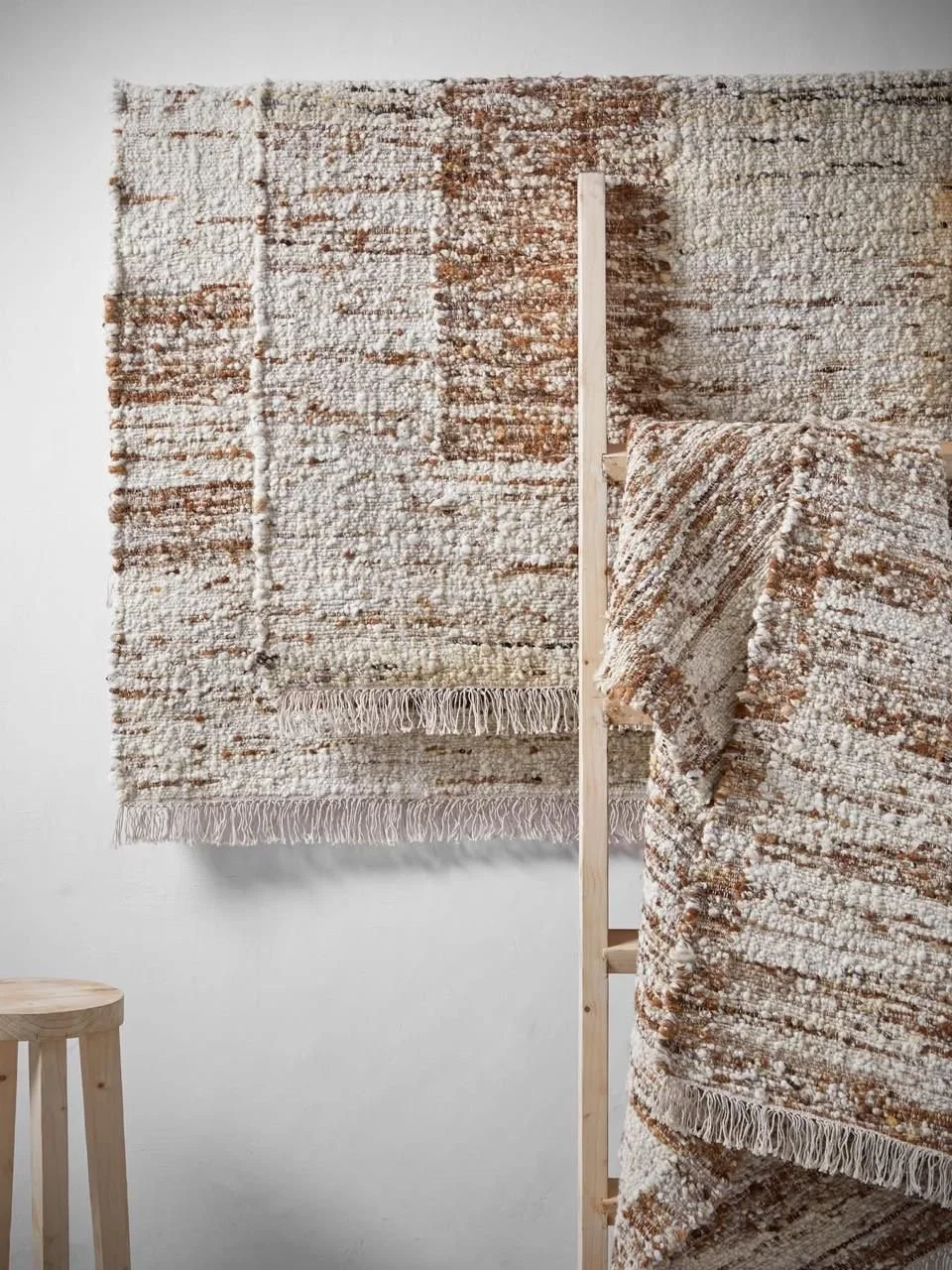 Two textured woven rugs with fringed edges in neutral and earth tones displayed on a wooden rack against a plain white wall, with a small wooden stool visible on the left.