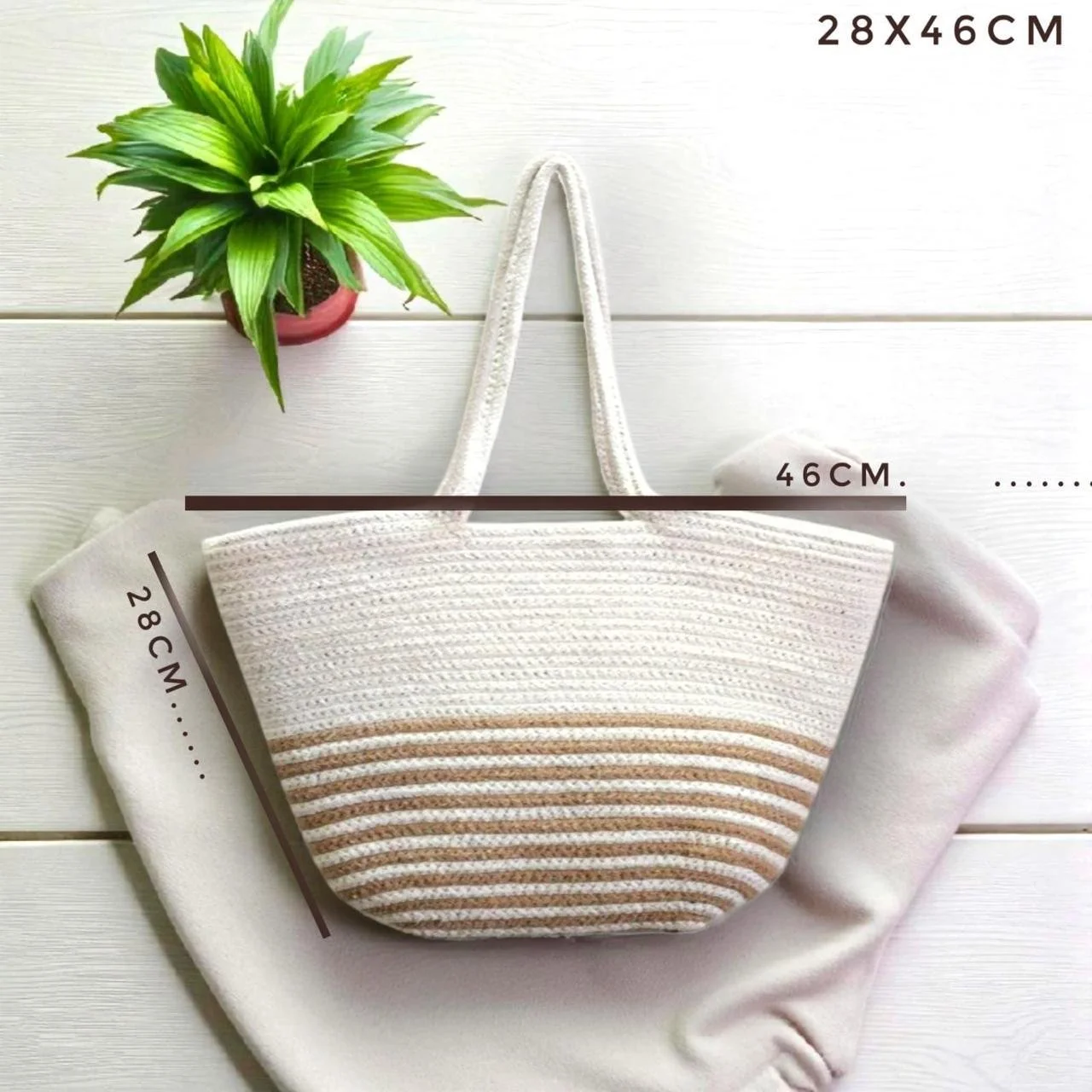 A striped woven tote bag with long handles, placed on a white cloth on a white wooden surface, with a small green potted plant nearby. The bag's dimensions are 28 cm in height, 46 cm in width, and 28 cm in depth.
