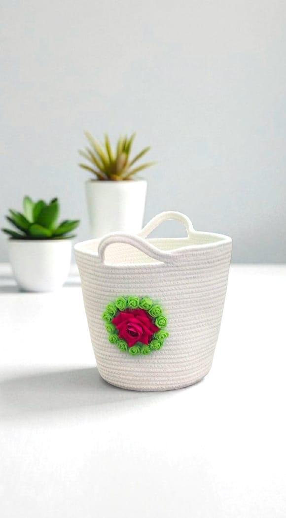 White woven basket with a pink rose and green ribbon decoration, placed on a white surface. Two potted succulents in white pots are in the background.