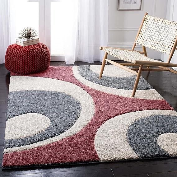 A geometric area rug with swirling shapes in white, gray, and red on a dark hardwood floor, a woven chair with wooden legs, and a red pouf with a small plant and books on top near a window with white curtains.