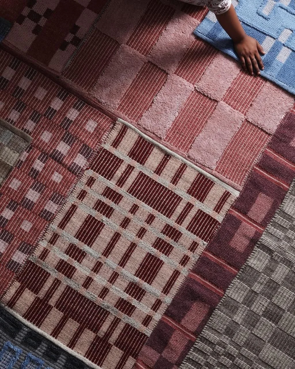 Close-up view of a person's hand on a blue and white rug, with various patterned rugs underneath on the floor.