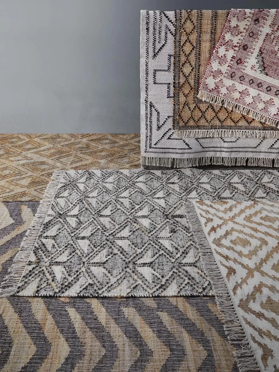 Multiple decorative rugs with various patterns and colors layered on top of each other, placed on a wooden floor against a plain wall.