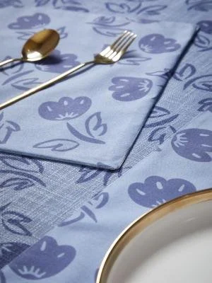 A table setting with a blue napkin and tablecloth featuring a floral pattern, a gold-rimmed white plate, and a spoon and fork placed on the napkin.