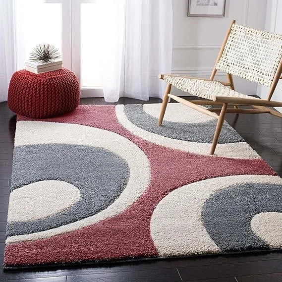 Living room corner with patterned rug, wicker chair, red pouf with books and decorative plant, white curtains, white wall, and framed artwork.