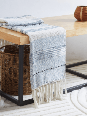 A woven throw blanket with fringe hanging over the edge of a wooden table, with a basket underneath and a textured rug on the floor.