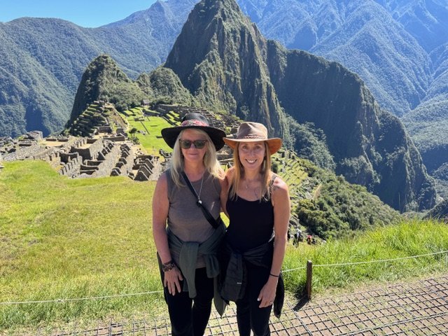 Two women wearing hats and sunglasses standing in front of Machu Picchu, an ancient Incan citadel in the mountains of Peru.