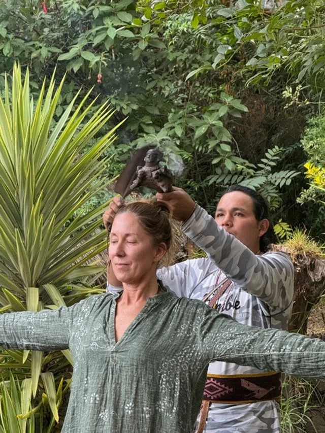 A woman with closed eyes and arms outstretched receives a prayer or blessing from a man holding a small statue above her head in a lush, green garden.