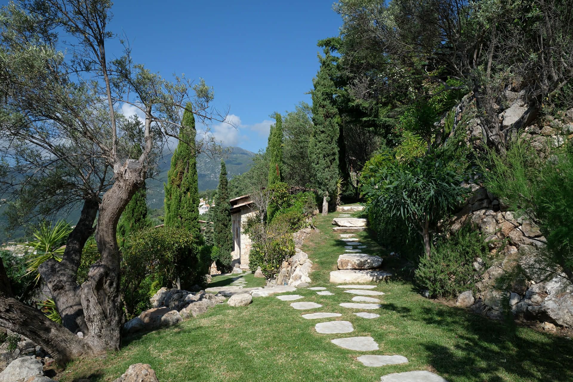 A stone pathway winds through a lush garden with tall trees and bushes on both sides. The sky is clear and blue with some clouds, and there are mountains in the background.