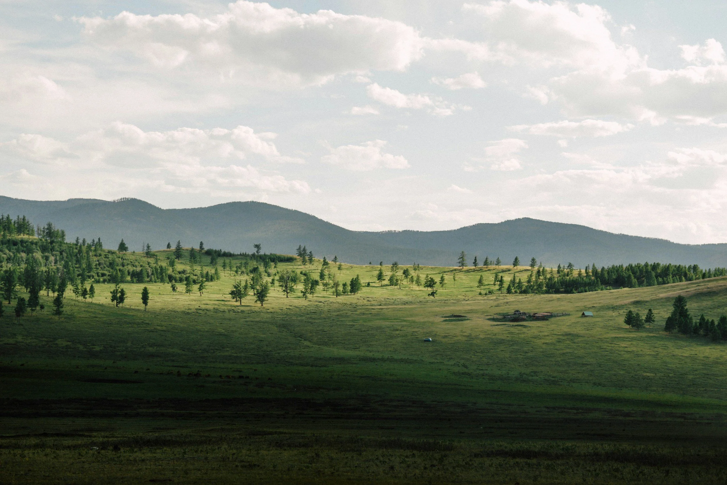 A landscape view of rolling green hills with scattered trees, distant mountains, and a partly cloudy sky.