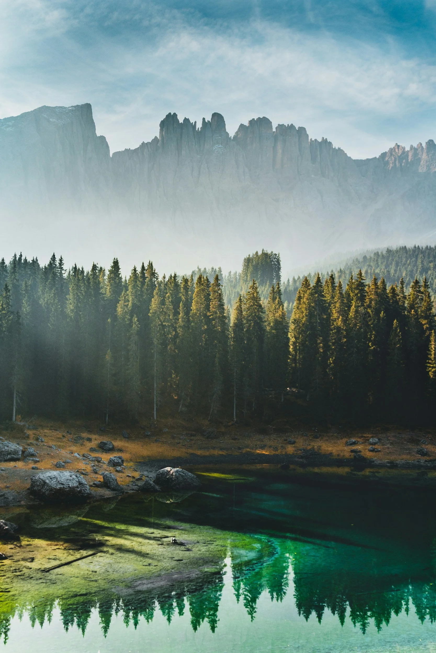 A mountain range with jagged peaks under a blue sky with wispy clouds. Dense evergreen forest in the foreground with a small body of water reflecting the trees.
