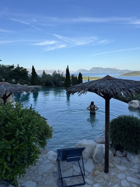 Person relaxing in a swimming pool with straw umbrellas, surrounded by greenery, overlooking a lake and mountains under a blue sky.