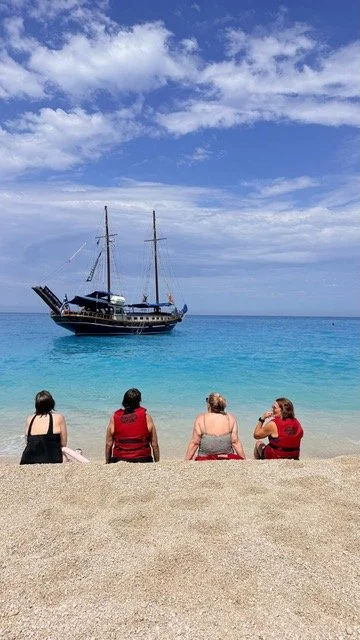 Four women sitting on a sandy beach facing the ocean, with a large sailboat anchored in the turquoise water under a partly cloudy sky.