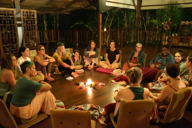 Group of people sitting in a circle around a small fire inside a wooden structure with tropical plants in the background.