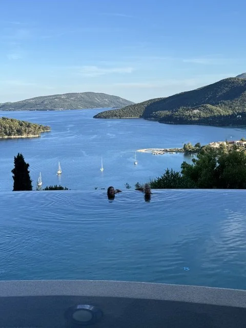 Two people swimming in an infinity pool overlooking a large lake with sailboats, surrounded by hills and a clear blue sky.