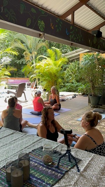 People practicing yoga on mats in a lush outdoor garden space with plants and trees, while others sit nearby at a table under a roofed patio.