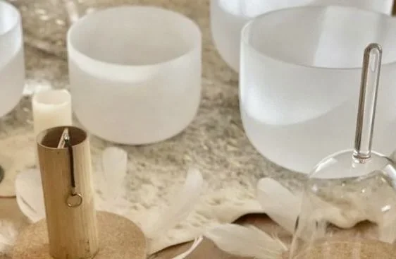 Various clear crystal singing bowls on a wooden surface, with a wooden mallet, surrounded by a white cloth and small white candles.