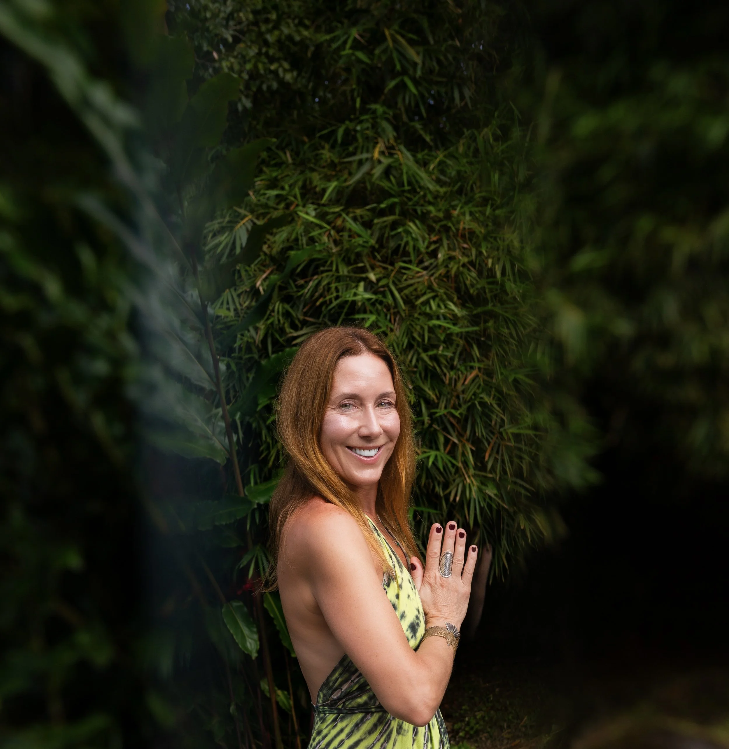A smiling woman with red hair in a yellow and green dress standing next to lush green bushes in an outdoor setting.