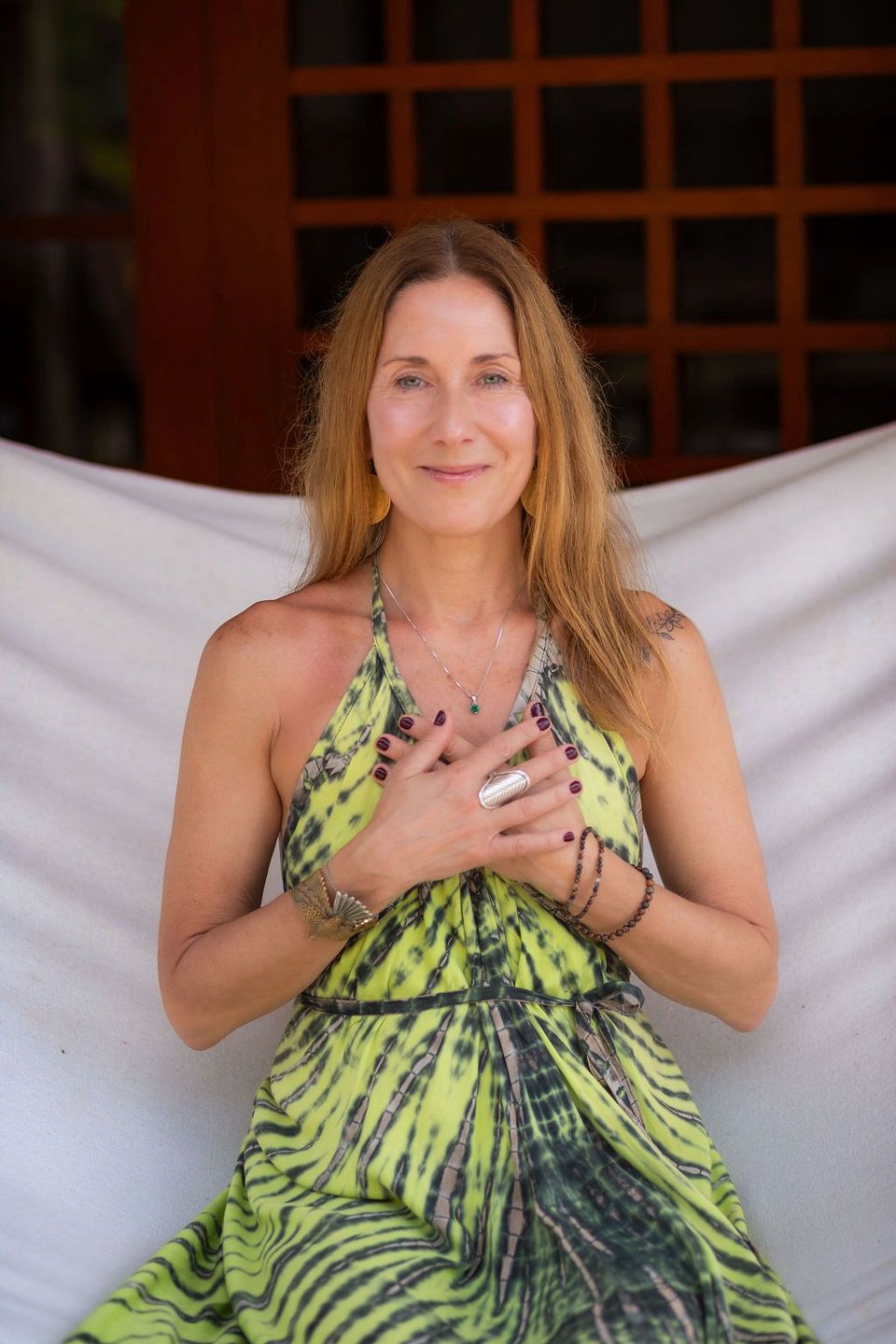 A woman with long reddish-brown hair and light skin, smiling with her hands on her chest, wearing a yellow and black patterned dress, jewelry including necklaces and rings, sitting in front of a wooden background and white sheet.