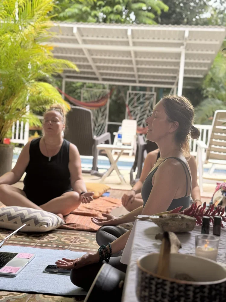 Three women practicing meditation outdoors on a patio, seated cross-legged on cushions, with a shaded backyard, potted plants, and outdoor furniture in the background.