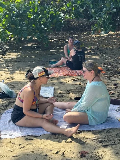 Two women sitting on beach towels talking, with a third woman and a man in the background near lush green foliage.