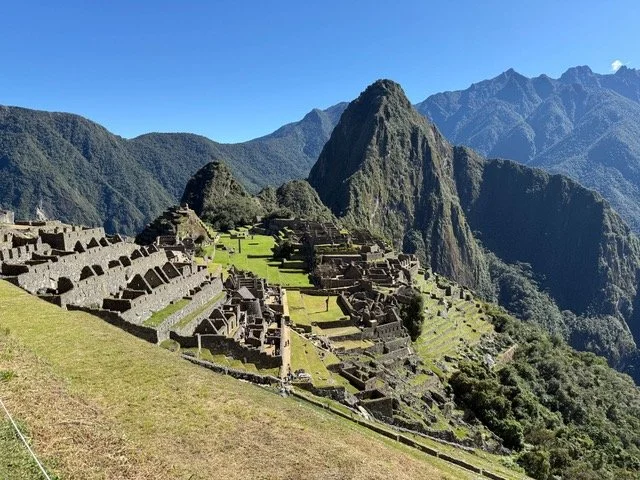 Ancient Incan ruins with terraced stone buildings on a hillside, surrounded by lush green mountains under a clear blue sky at Machu Picchu.