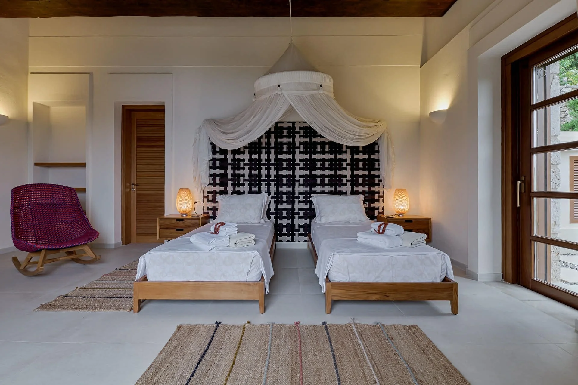 A bedroom with two single beds, each with white bedding, pillows, and towels, separated by a nightstand with a lamp. A decorative canopy hangs above the beds, and there is a large window on the right side with wooden framing. A red wicker rocking chair is on the left side, and a striped rug lies on the tile floor.