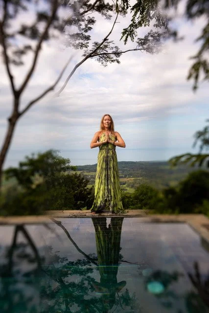 A woman in a green dress standing on a platform outdoors, with her hands in a prayer position, surrounded by trees and overlooking a scenic landscape.