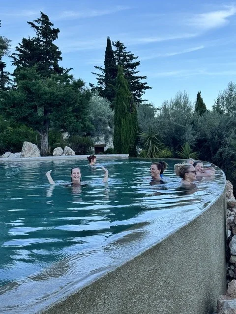 Group of women swimming and relaxing in an outdoor pool surrounded by trees and greenery.
