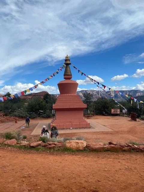 A large red Buddhist stupa with prayer flags draped around it, set in a desert landscape with mountains in the background.