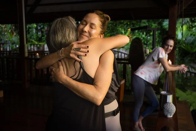 Two women hugging passionately, with a woman in the background clapping and smiling in an outdoor setting surrounded by lush green foliage.