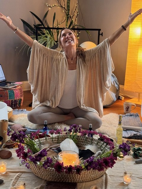 Woman sitting cross-legged on the floor with arms raised, smiling, in a cozy, well-lit room decorated with plants, candles, and various spiritual items.