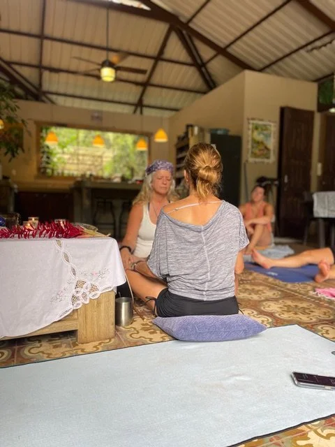 Women participating in a group meditation or yoga class inside a rustic, open-air room with high ceilings.