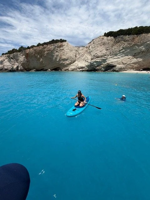 A woman paddleboarding on blue water near white cliffs with a person swimming nearby.