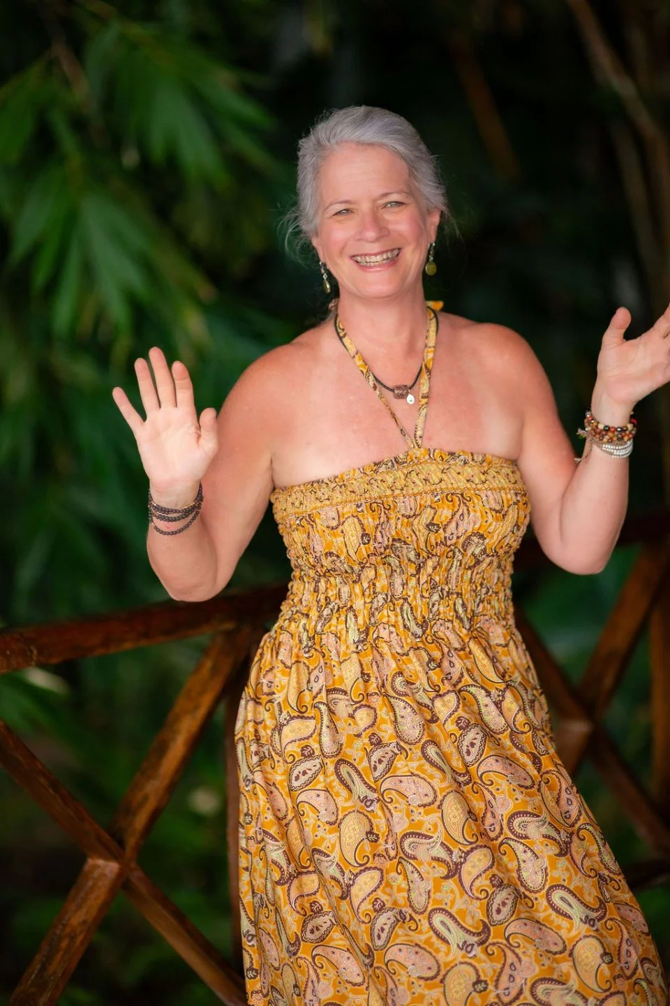 A smiling older woman with gray hair wearing a yellow paisley strapless dress and jewelry, standing outdoors in front of green foliage, waving with both hands.