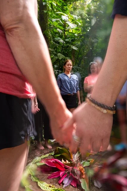 Two people holding hands over a floral arrangement in a jungle setting, with at least two women observing in the background.