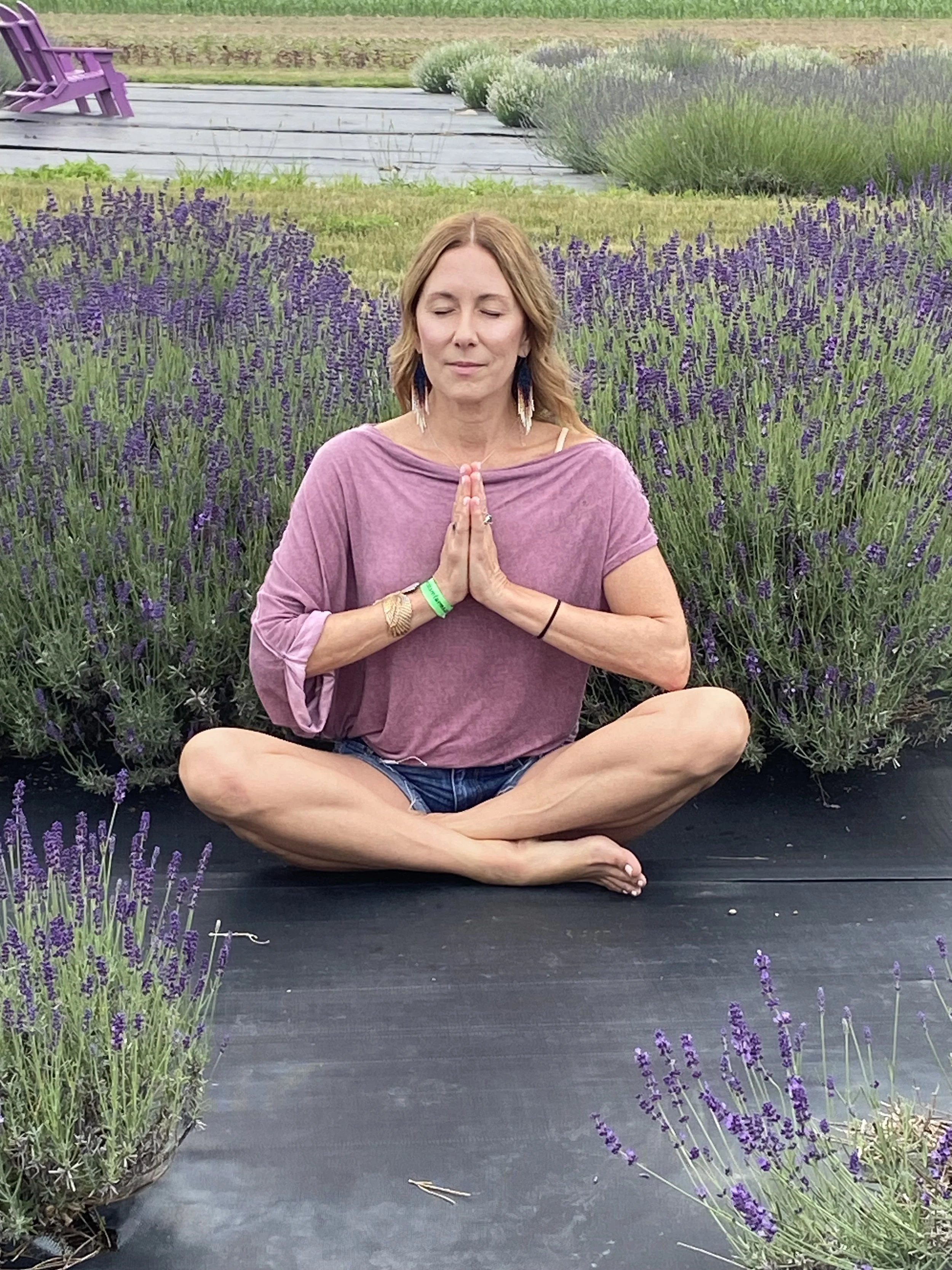 A woman practicing meditation or yoga in a lavender field, sitting cross-legged on a black mat with her hands in a prayer position, eyes closed, wearing a purple top and denim shorts.