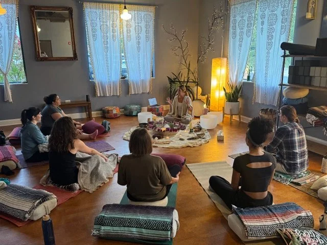 A group of people participating in a meditation or yoga session in a cozy, well-lit room with large windows, curtains, and decorative elements, including a lamp, plants, and cushions.